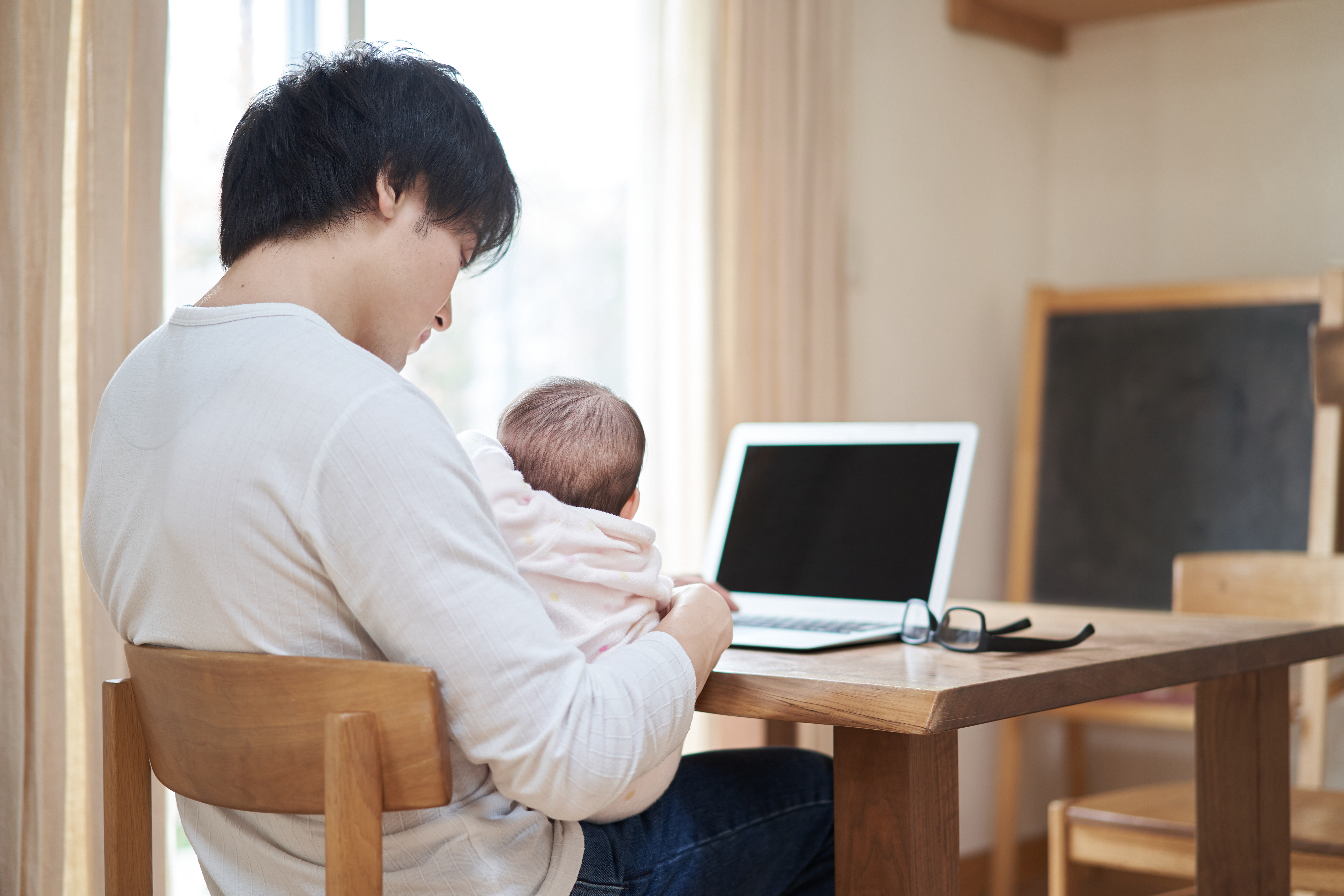 Photo from behind of a black haired man holding a young baby on his lap, while sitting at a wooden table at home in the daytime and typing on a laptop.